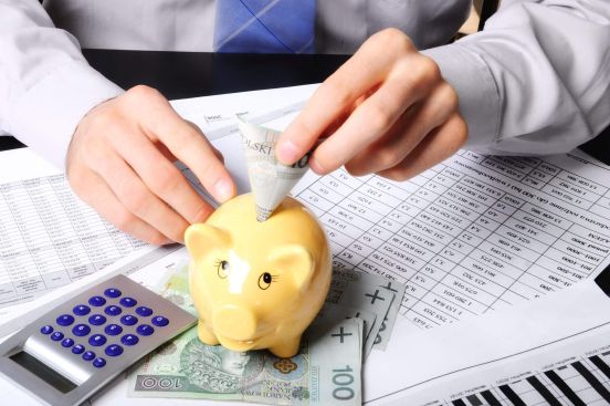 Ręka wsuwająca banknot do świnki-skarbonki stojącej obok kalkulatora